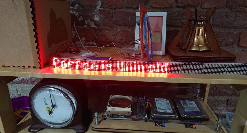 A photo of a shelf in DoES Liverpool showing an LED matrix strip informing us that the Coffee is 4 minutes old. The font used is quite compact. Around this are various technological devices including a server made of just components mounted on board and acrylic sheet, a clock showing locations around the edge and an internet connected bell.