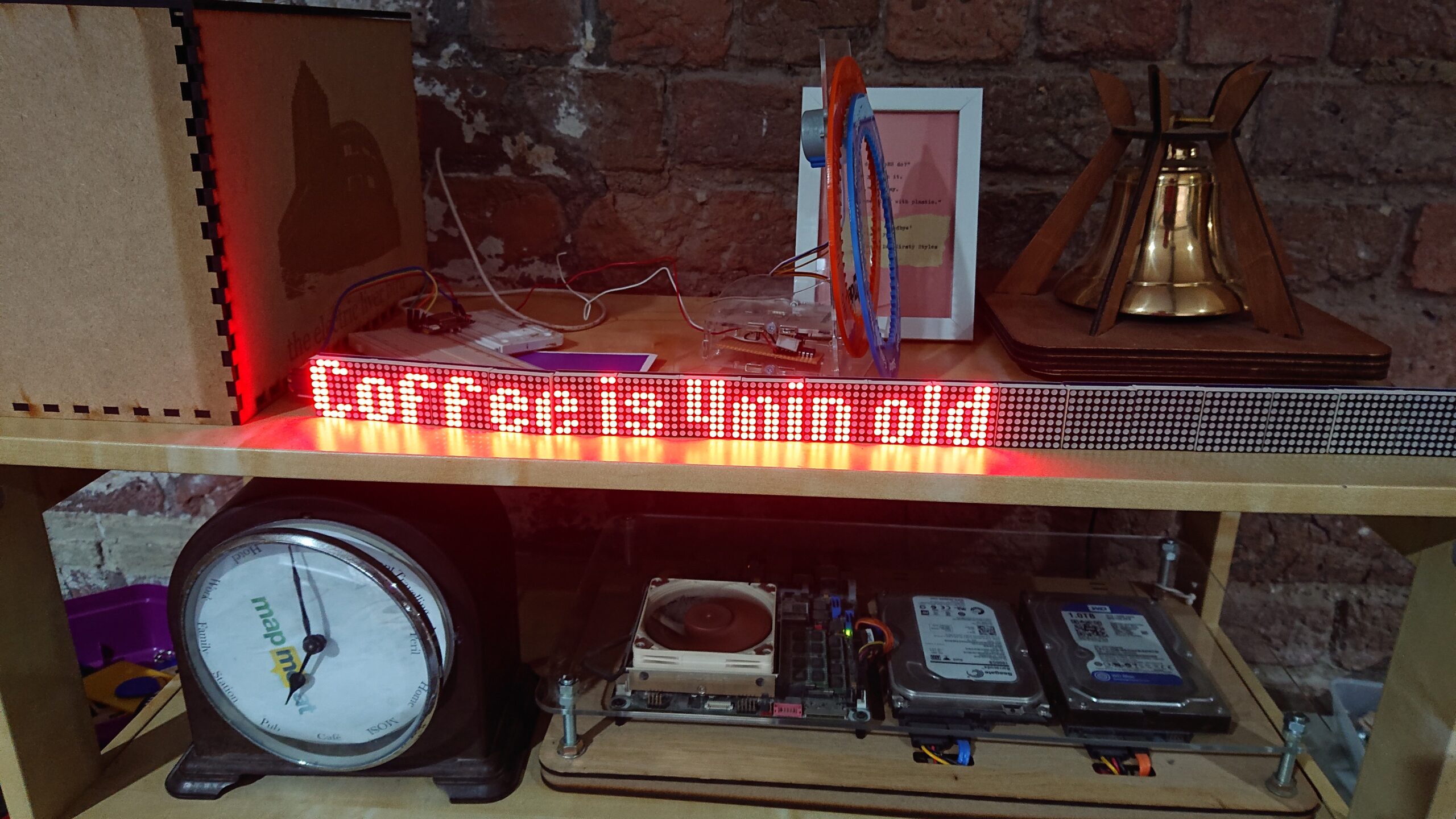 A photo of a shelf in DoES Liverpool showing an LED matrix strip informing us that the Coffee is 4 minutes old. The font used is quite compact. Around this are various technological devices including a server made of just components mounted on board and acrylic sheet, a clock showing locations around the edge and an internet connected bell.