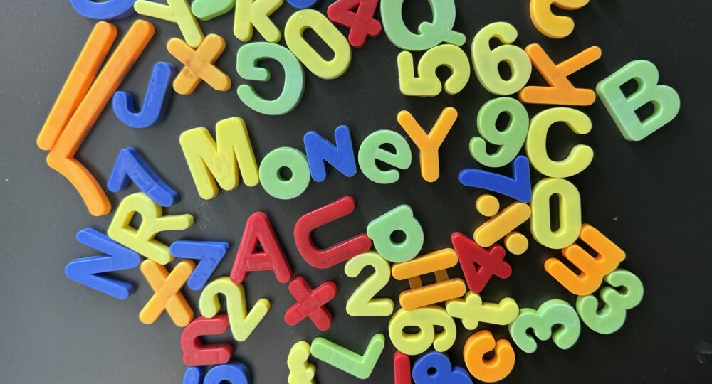 A selection of multi-coloured plastic letters attached to a fridge. The letters are mostly randomly places although the word "Money" can be read in the middle and "2+2=4" is also visible.