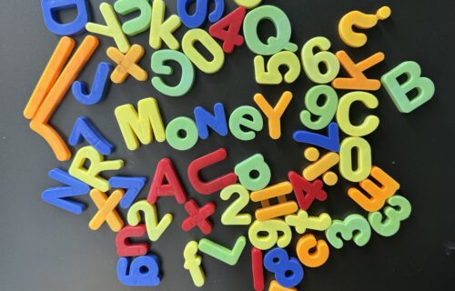 A selection of multi-coloured plastic letters attached to a fridge. The letters are mostly randomly places although the word "Money" can be read in the middle and "2+2=4" is also visible.