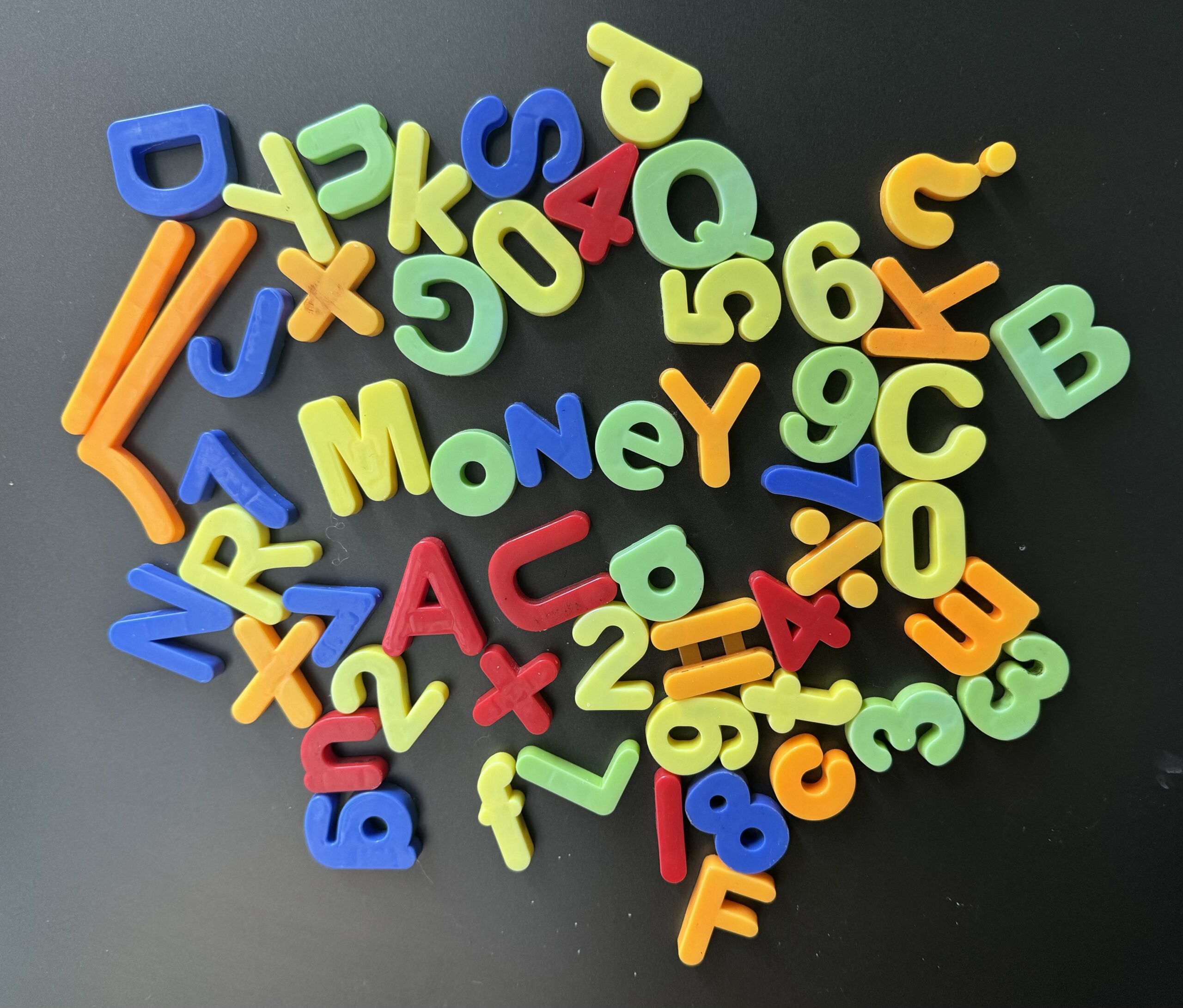 A selection of multi-coloured plastic letters attached to a fridge. The letters are mostly randomly places although the word "Money" can be read in the middle and "2+2=4" is also visible.