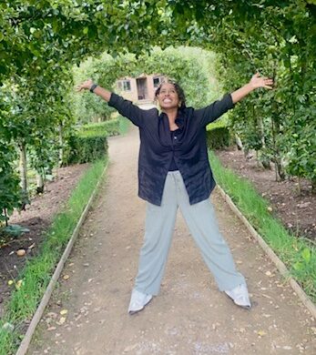 Sahra wearing a black top and grey leggings, smiling, on a garden path