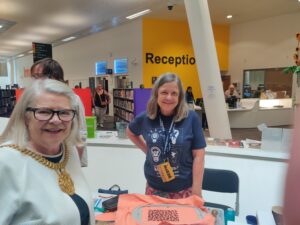 Lord Mayor Barbara in front of a desk, on which is a frame from the embroidery machine. Jackie is standing behind the desk wearing a MakeFest t shirt